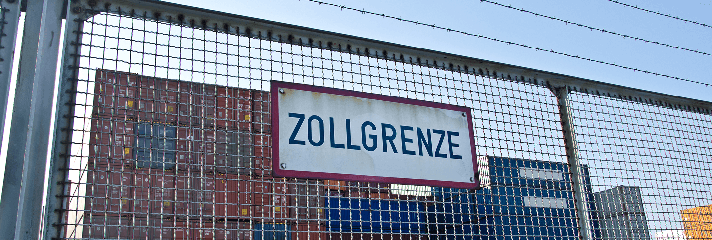 Metallschild mit der Aufschrift Zollgrenze. Das Schild hängt an einem mit Stacheldraht gesicherten Zaun. Dahinter sieht man sich stapelnde Container